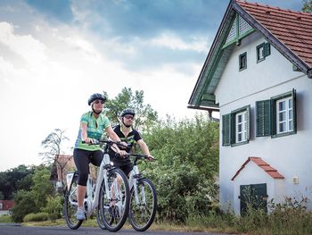 Zwei Radfahrer fahren auf einem Weg vorbei an einem traditionellen Haus mit grünen Fensterläden, umgeben von Bäumen und einer idyllischen Dorfkulisse.