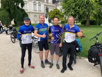 Four cyclists in matching jerseys pose with thumbs up in a park, with bicycles and greenery in the background.