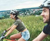 Eine junge Frau und ein Mann auf Fahrrädern fahren durch eine grüne Landschaft, im Hintergrund eine Stadtansicht und blauer Himmel.
