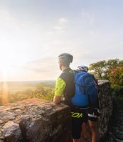 Zwei Radfahrer genießen den Sonnenuntergang an einem Aussichtspunkt mit weitem Blick auf eine grüne Landschaft.