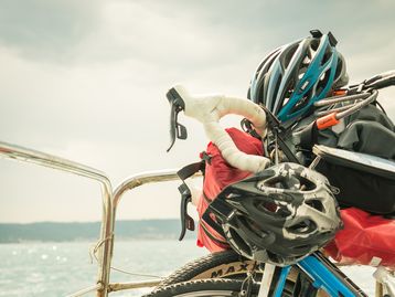 Nahaufnahme eines Fahrrads mit Helm und Packtaschen, das auf einer Fähre transportiert wird, mit Blick auf das Wasser und eine entfernte Küstenlinie.