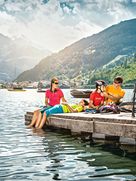 Eine Familie von vier Personen entspannt auf einem hölzernen Steg am See, umgeben von malerischen Bergen. Ein Erwachsener hilft einem Kind beim Einstellen des Helms, während das andere Kind am Steg liegt und die Füße im Wasser baumeln lässt. Fahrräder und Helme liegen neben ihnen, während Boote auf dem See treiben.