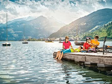 Eine Familie von vier Personen entspannt auf einem hölzernen Steg am See, umgeben von malerischen Bergen. Ein Erwachsener hilft einem Kind beim Einstellen des Helms, während das andere Kind am Steg liegt und die Füße im Wasser baumeln lässt. Fahrräder und Helme liegen neben ihnen, während Boote auf dem See treiben.