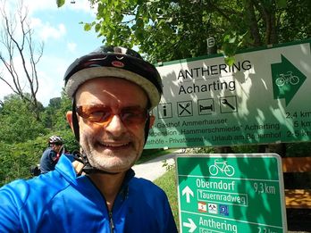 Hannes Friedrich in a blue EuroVelo jersey smiles in front of green directional signs and a large information board on a sunny day.