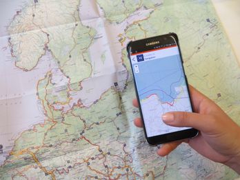 Eine Hand hält ein Smartphone mit einer geöffneten EuroVelo-App, die eine Routenkarte anzeigt. Im Hintergrund liegt eine detaillierte Papierkarte mit markierten EuroVelo-Routen.