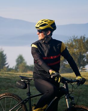 Ein Radfahrer in schwarz-gelber Ausrüstung hält an, um eine in Nebel gehüllte Berglandschaft zu bewundern. Dabei stützt er sich mit den Händen auf seinem Fahrrad ab und blickt zur Seite.