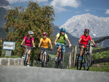Vier Radfahrer*innen in farbenfroher Kleidung radeln lächelnd durch eine ländliche Umgebung mit Obstbäumen und hohen Bergen im Hintergrund. Ein Schild mit der Aufschrift „Almdorf“ ist am Straßenrand zu sehen, während die Gruppe gemeinsam die Landschaft genießt.