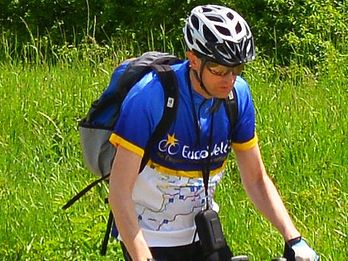 Andrzej Felczak in EuroVelo-Trikot fährt mit einem roten Fahrrad durch eine grüne Landschaft, ausgestattet mit Helm, Sonnenbrille und einem grauen Rucksack.