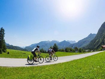Zwei Radfahrer auf einer asphaltierten Straße, umgeben von grünen Wiesen und beeindruckendem Alpenpanorama bei strahlendem Sonnenschein.