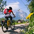 Ein Radfahrer mit rotem Rucksack fährt auf einem Feldweg mit Blick auf einen majestätischen Berg und eine idyllische Landschaft.