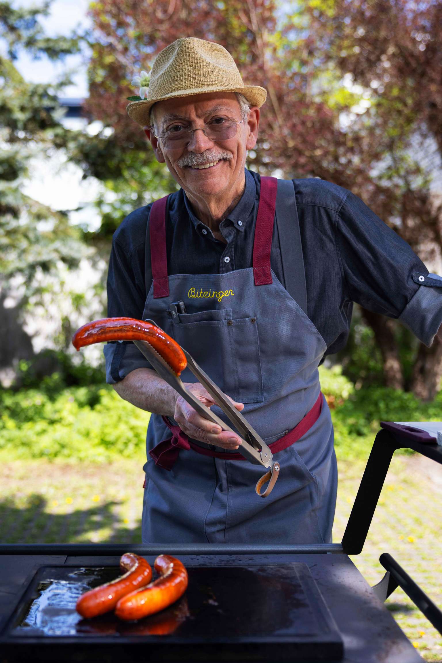 A smiling older man in a straw hat grills sausages outdoors, holding tongs and wearing a gray apron with red straps.