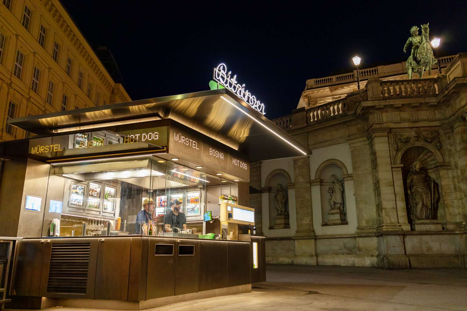 A modern nighttime food stand in a historic city setting, offering sausages and hot dogs, with an illuminated sign and statues in the background.