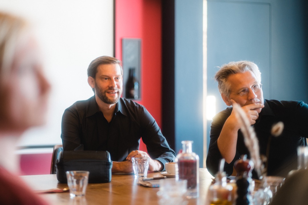 Zwei Männer in schwarzen Hemden sitzen am Tisch, dahinter eine rote und blaue Wand.