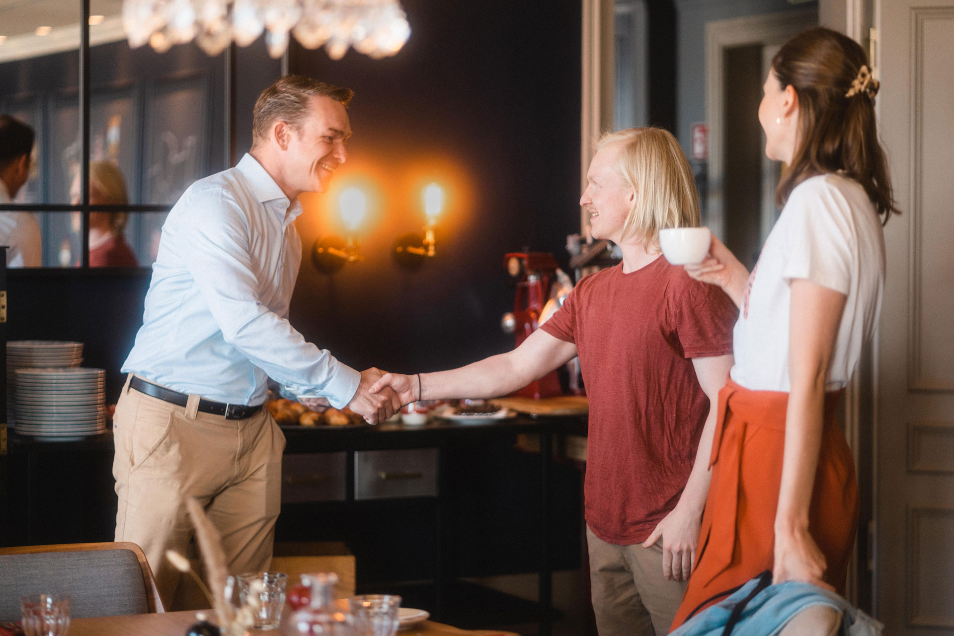 Zwei Männer begrüßen sich mit Handschlag, daneben eine Frau mit einer Kaffeetasse.