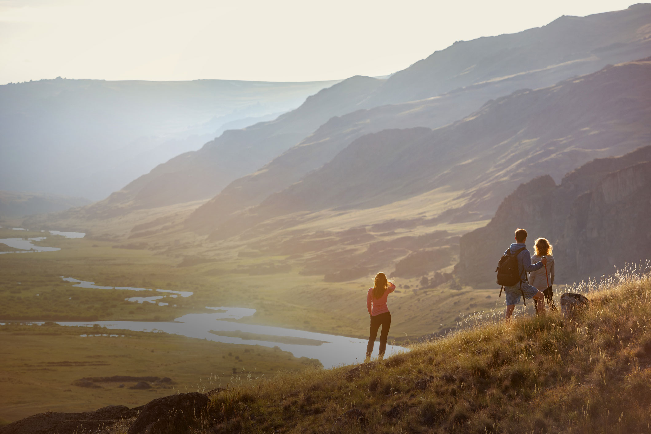 Drei Personen stehen auf einem Berg und blicken ins Tal mit Fluss.