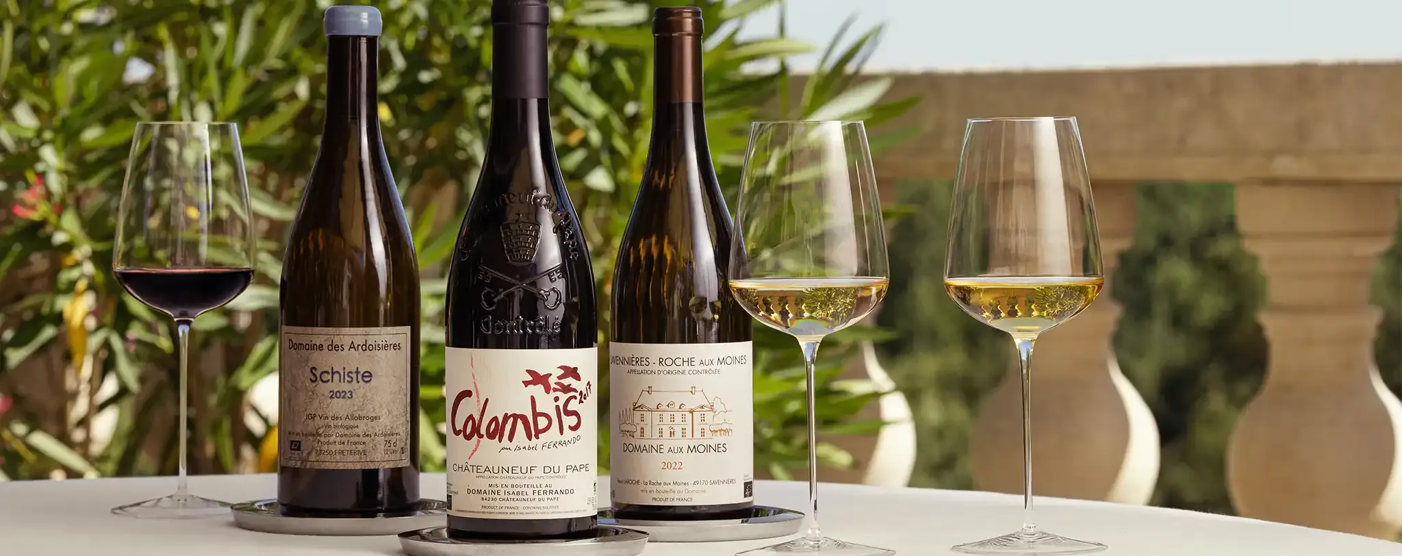 Trois bouteilles de vin et trois verres sur une table à l'extérieur, avec un arrière-plan feuillu et une balustrade en pierre.