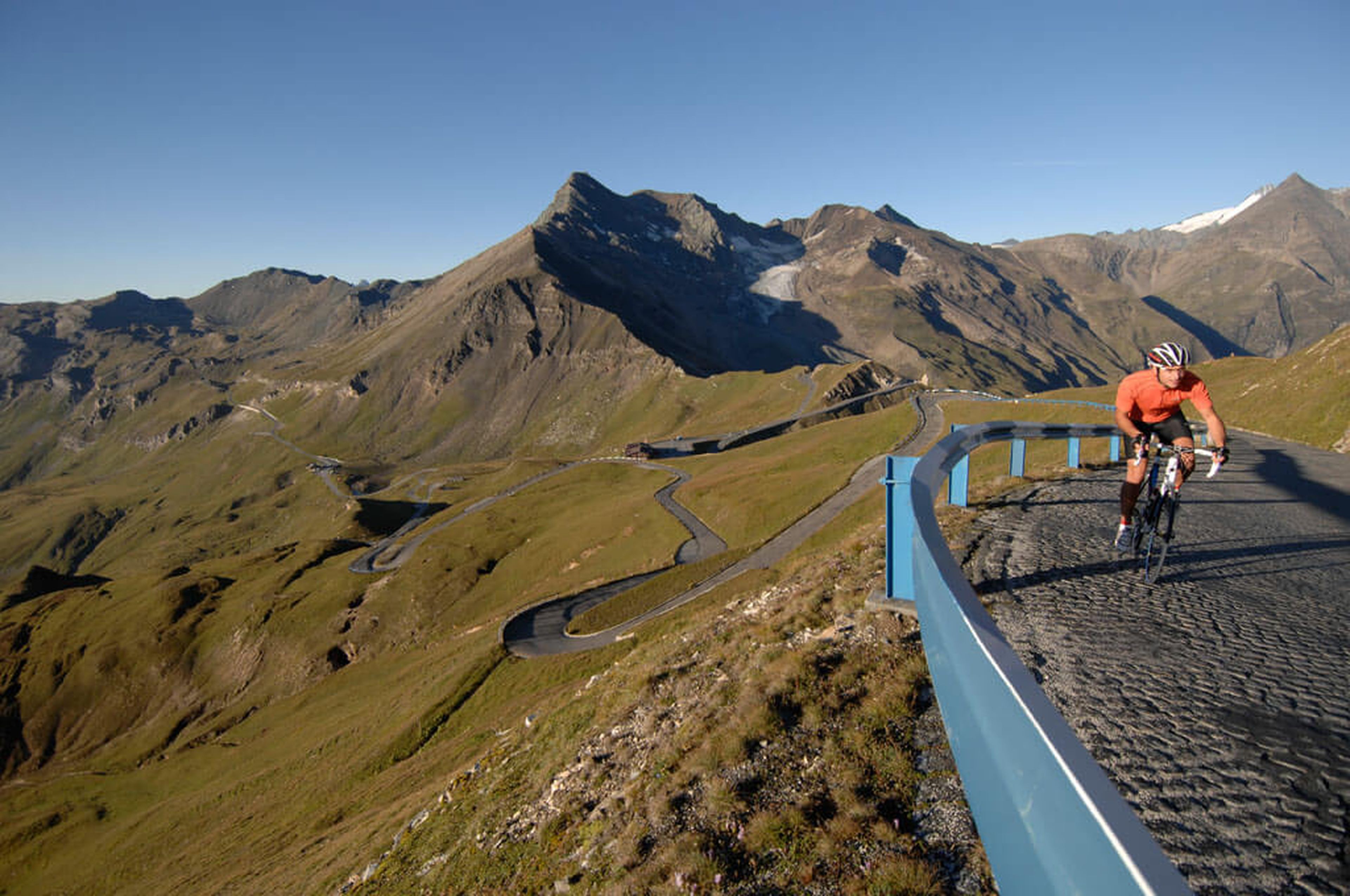 Radfahrer im orangefarbenen Hemd auf einer kurvenreichen Bergstraße mit schroffen Gipfeln und klarem blauen Himmel im Hintergrund.