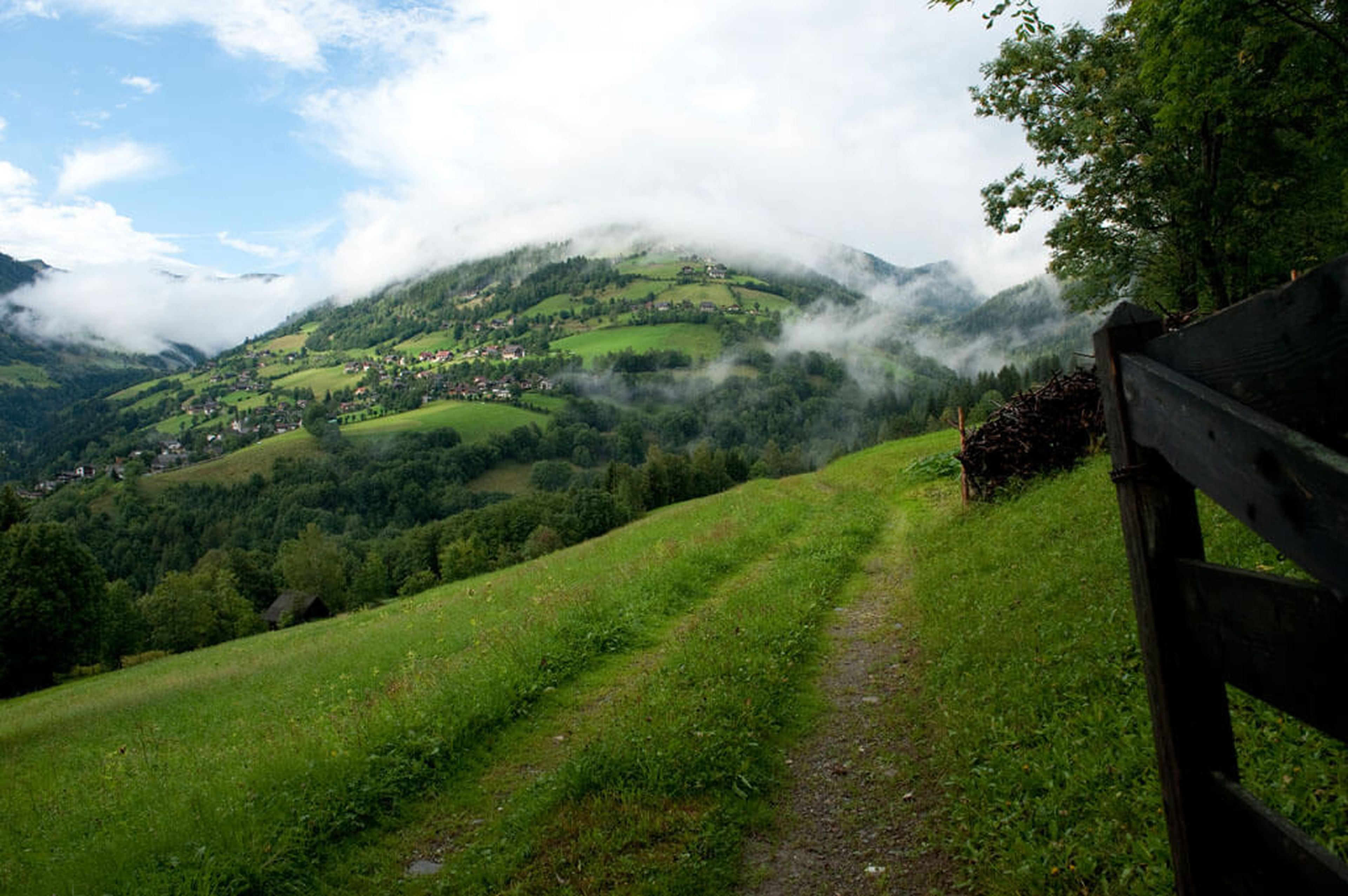 Ein üppig grüner Pfad am Hang führt zu einem weit entfernten Dorf, eingebettet in nebelverhangene, baumbewachsene Berge unter einem teilweise bewölkten Himmel.