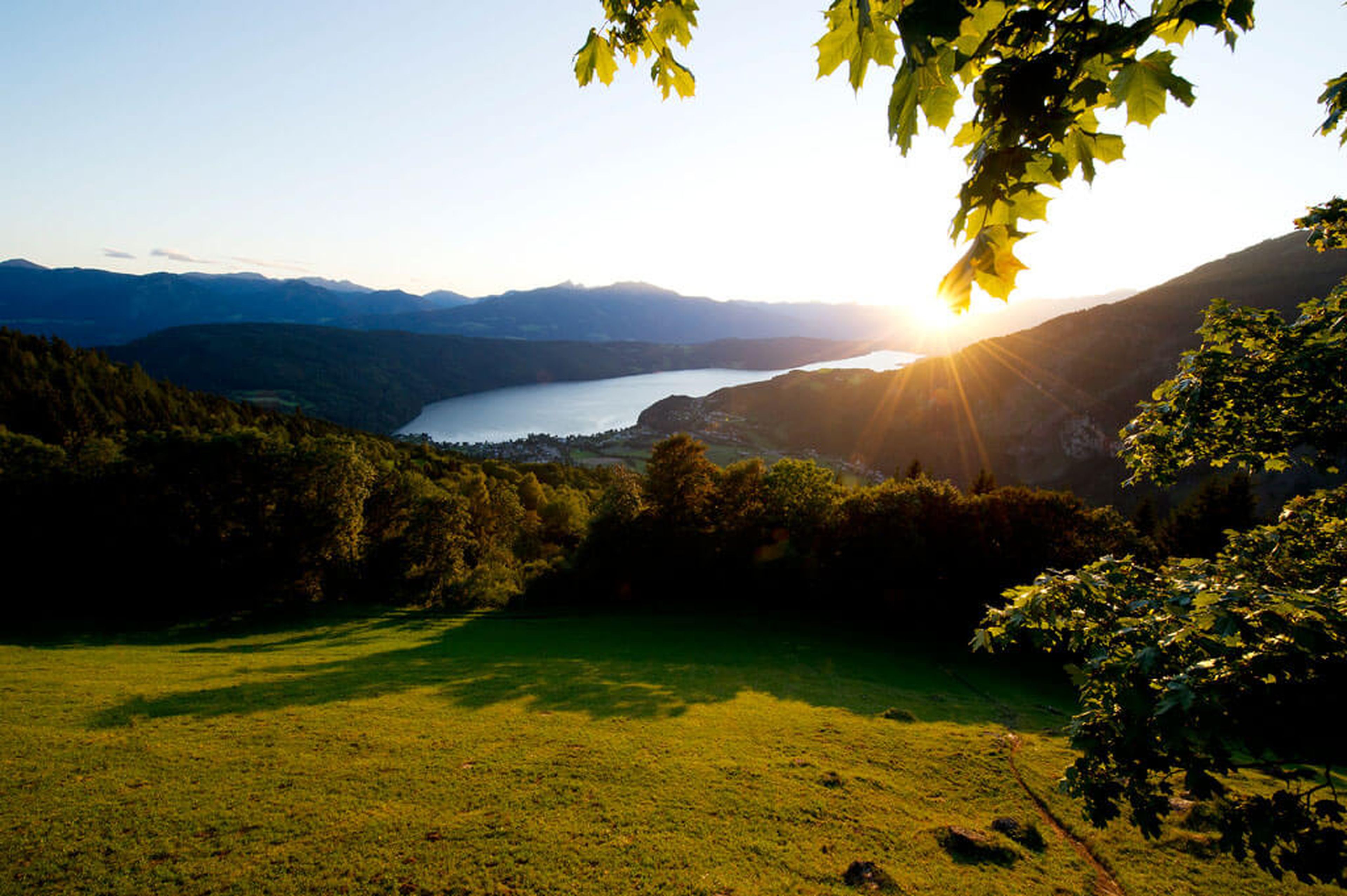 Sonnenuntergang über einem ruhigen See, umgeben von üppig grünen Hügeln und Bäumen, mit fernen Bergen unter einem klaren blauen Himmel.
