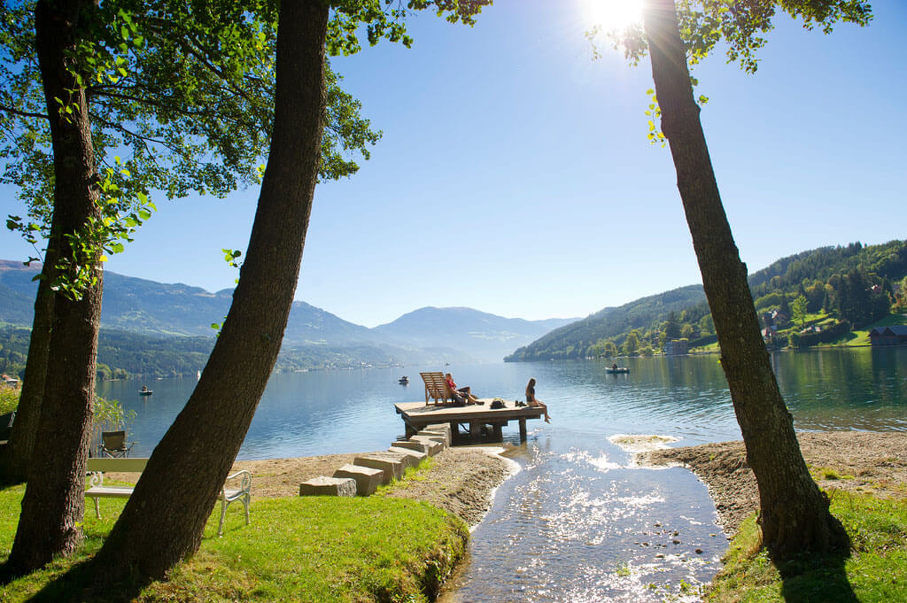 Eine ruhige Seeszene mit zwei Personen, die auf einem hölzernen Steg sitzen, umgeben von Bäumen und Bergen unter einem klaren blauen Himmel.