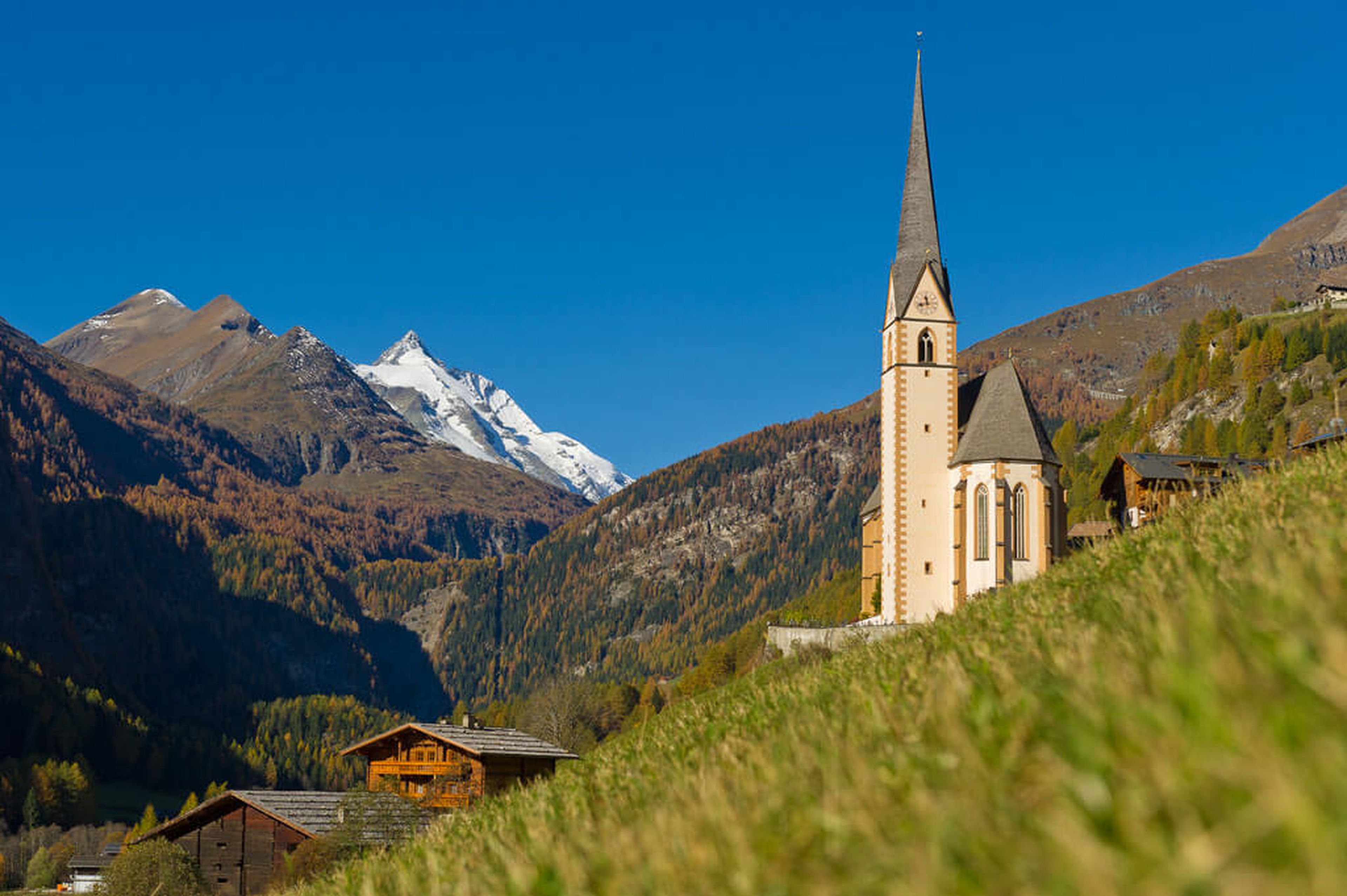 Eine hohe Kirche mit einem spitzen Turm steht auf einem grasbewachsenen Hügel, umgeben von Holzhäusern, mit schneebedeckten Bergen im Hintergrund unter einem klaren blauen Himmel.
