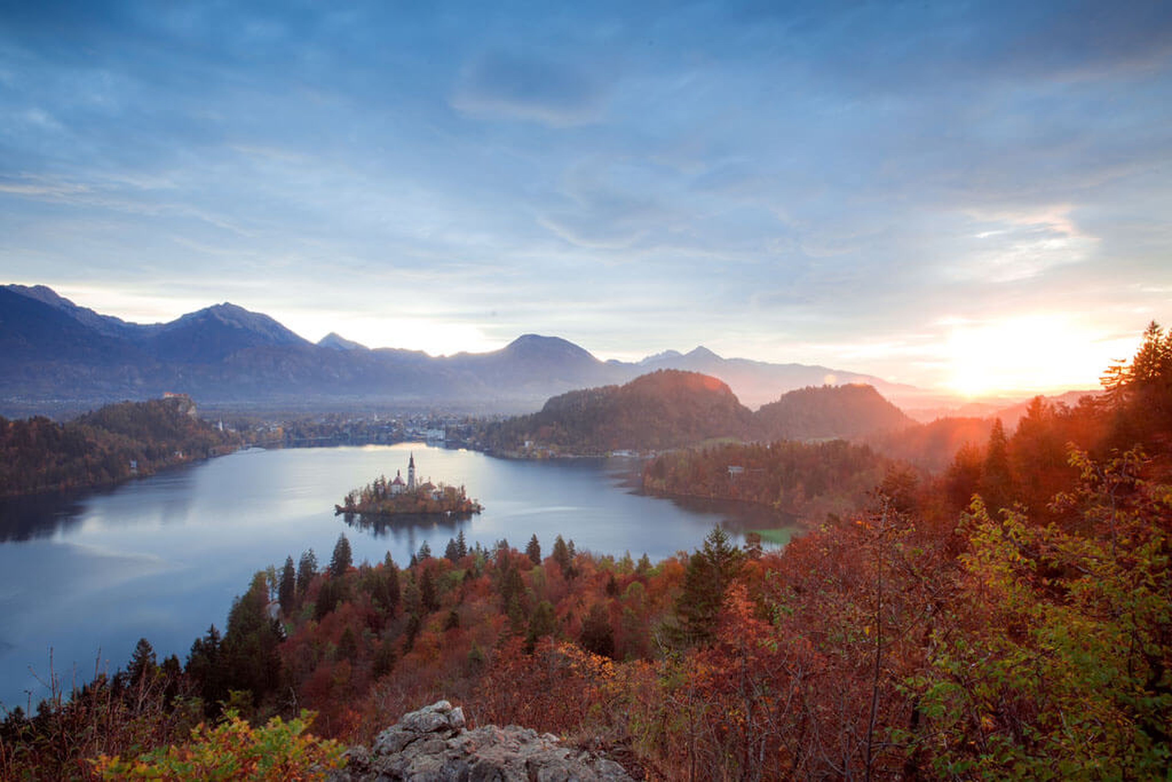 Ein ruhiger See mit einer kleinen Insel und einer Kirche, umgeben von herbstlichen Bäumen und Bergen unter einem strahlenden Sonnenaufgangshimmel.