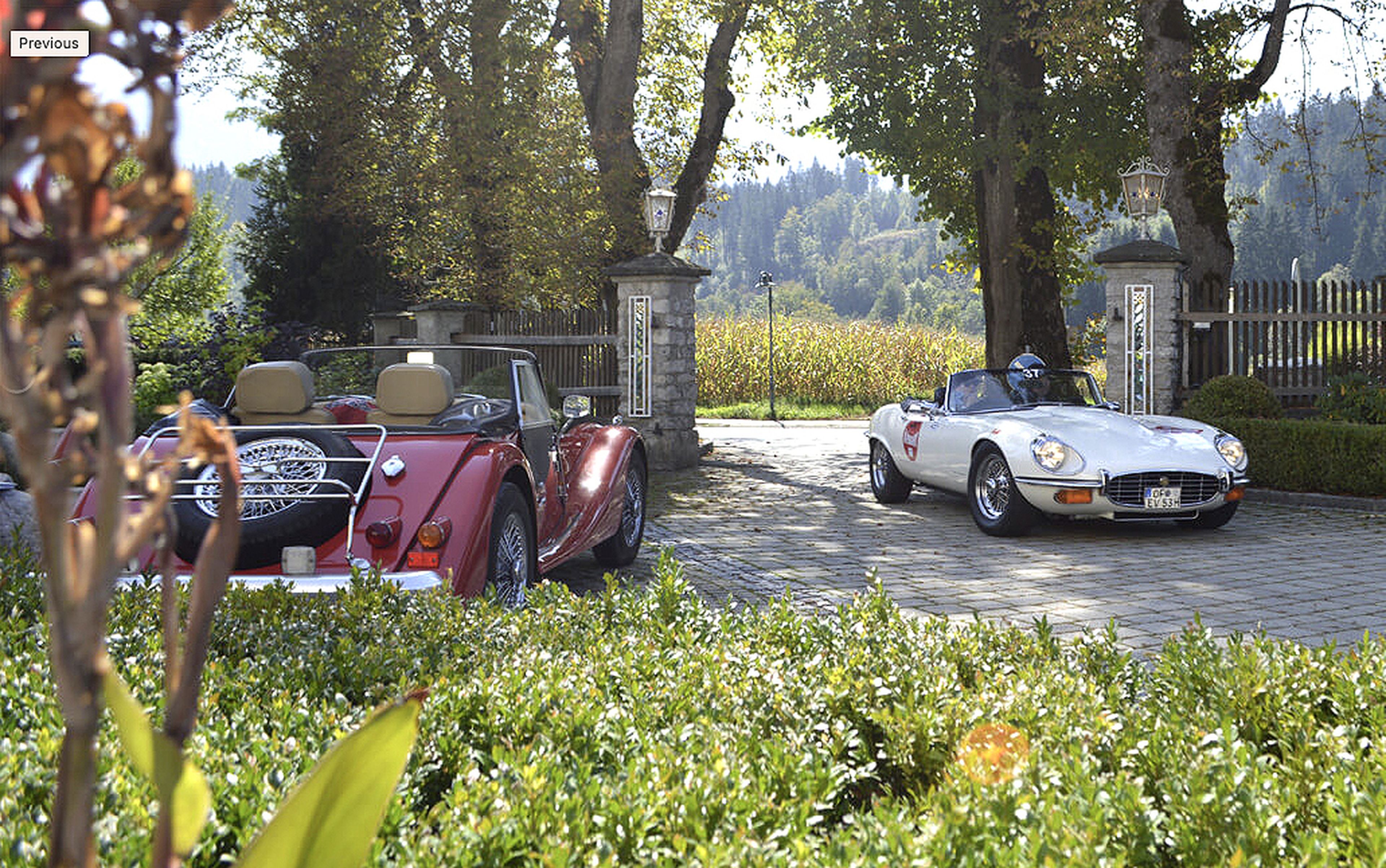 Zwei Oldtimer-Cabrios, geparkt auf einer kopfsteingepflasterten Auffahrt, umgeben von Grün, Bäumen und einem malerischen Hintergrund aus Hügeln und Feldern.