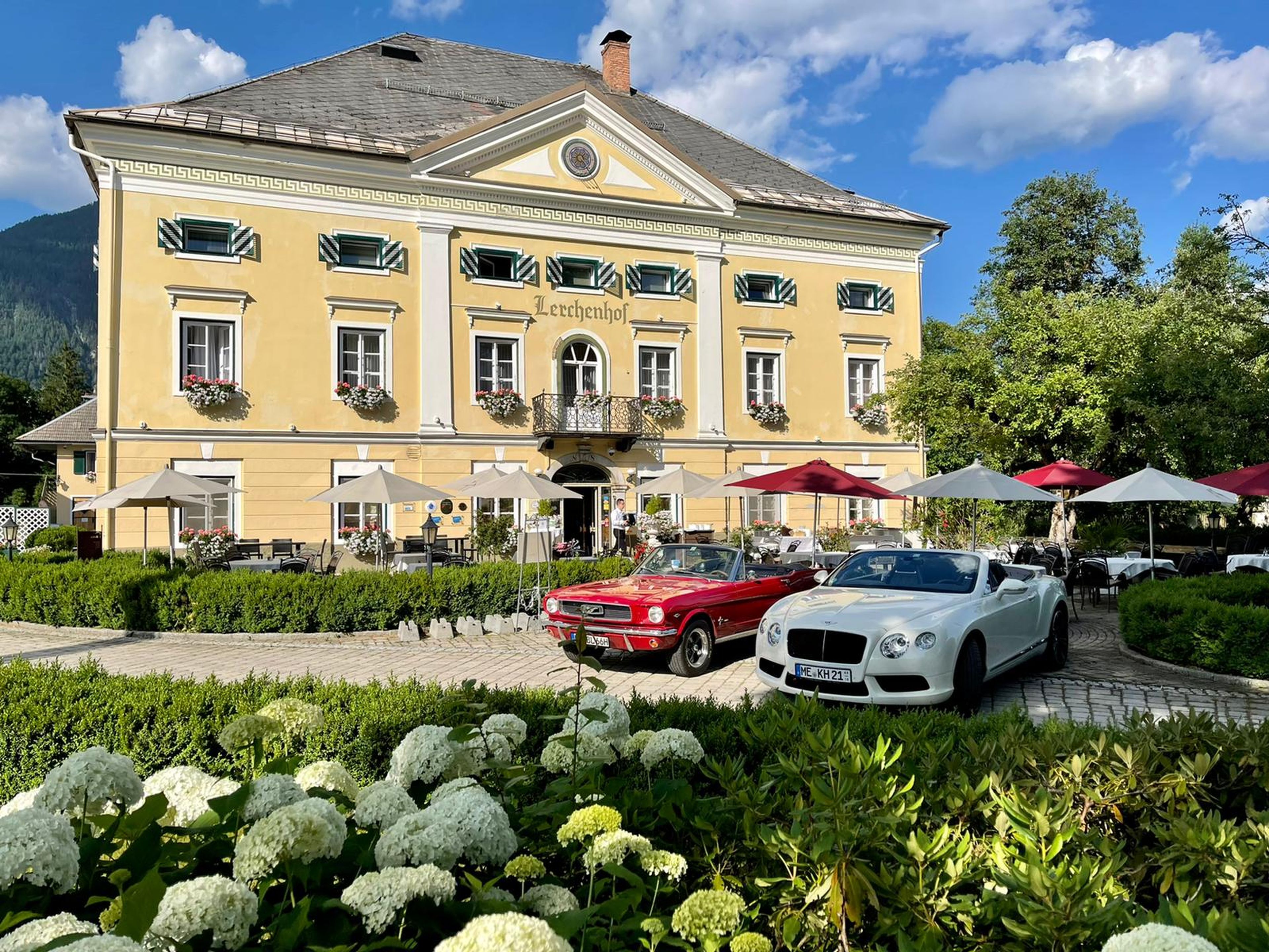 Schloss Lerchenhof – ein schönes historisches Gebäude, davor zwei geparkte Oldtimer, umgeben von üppigen Gärten und Sitzgelegenheiten im Freien unter Sonnenschirmen.