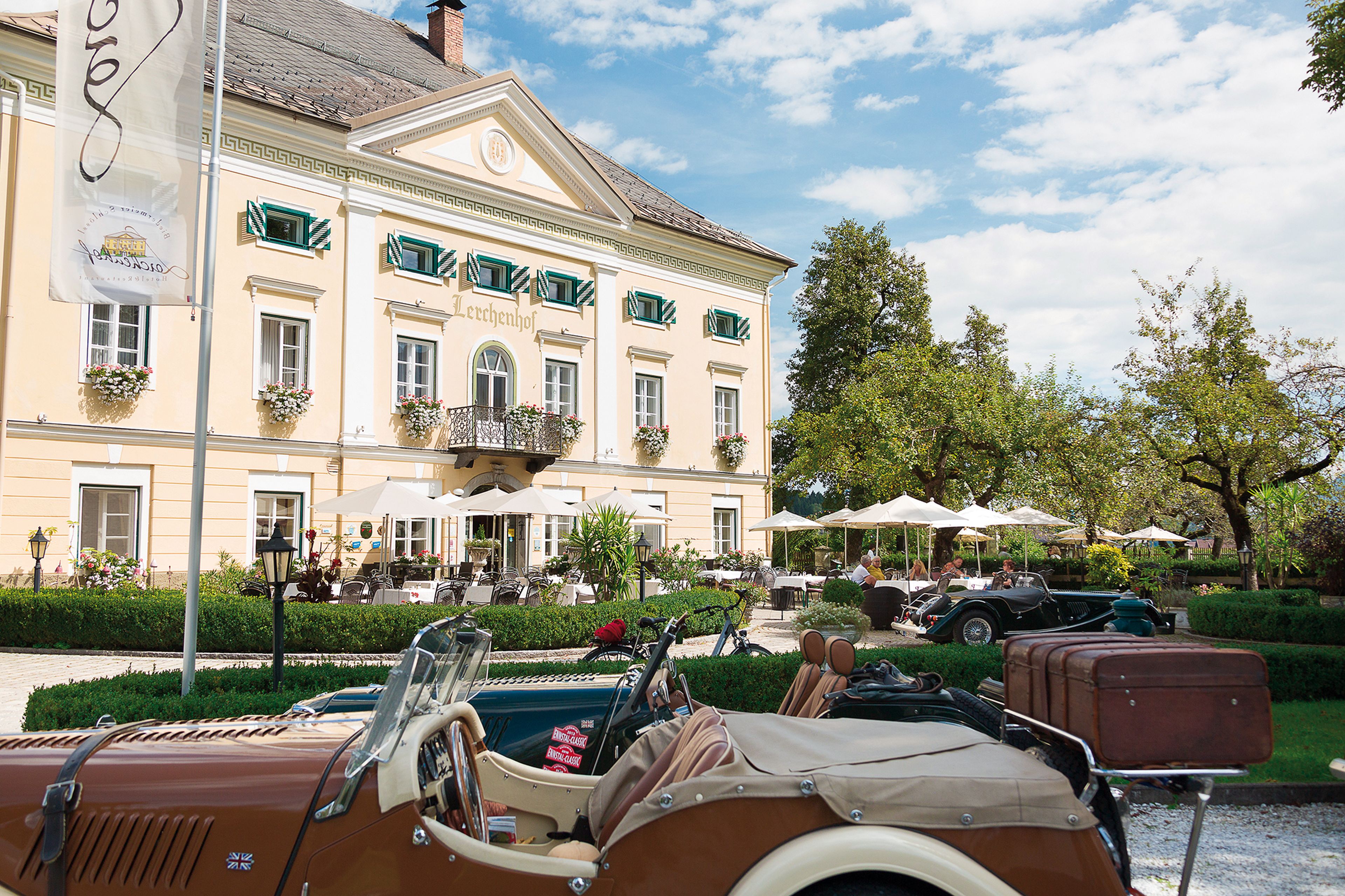 Oldtimer, die vor dem Schloss Lerchenhof geparkt sind, umgeben von Bäumen und Sitzgelegenheiten im Freien unter Sonnenschirmen.