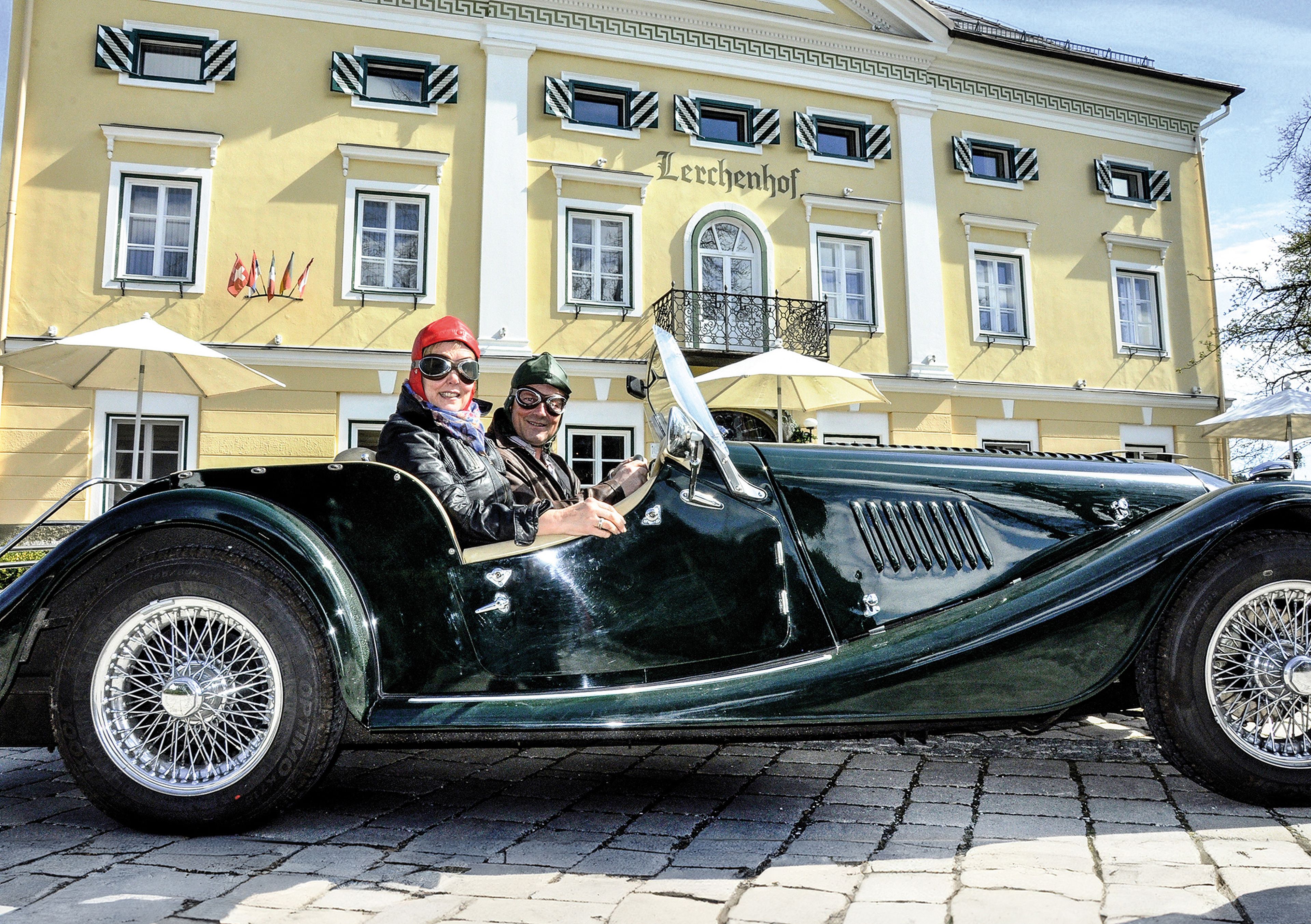 Zwei Personen in Oldtimerkleidung sitzen in einem Oldtimer, der vor einem gelben Gebäude mit der Aufschrift „Leccchenhof“ geparkt ist.
