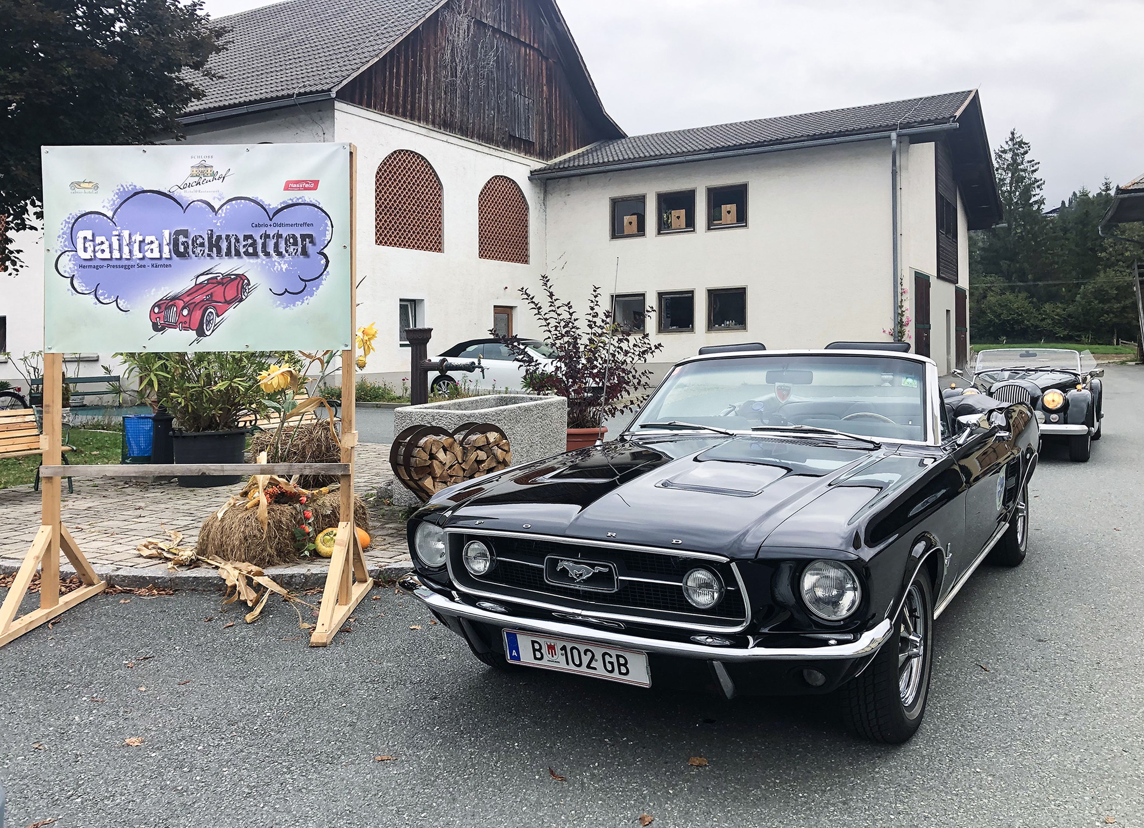 Ein klassisches schwarzes Cabrio parkt in der Nähe eines Schildes mit der Aufschrift „Gailtal Geknatter“, in dessen Nähe sich ein rustikales Gebäude und herbstliche Dekorationen befinden.