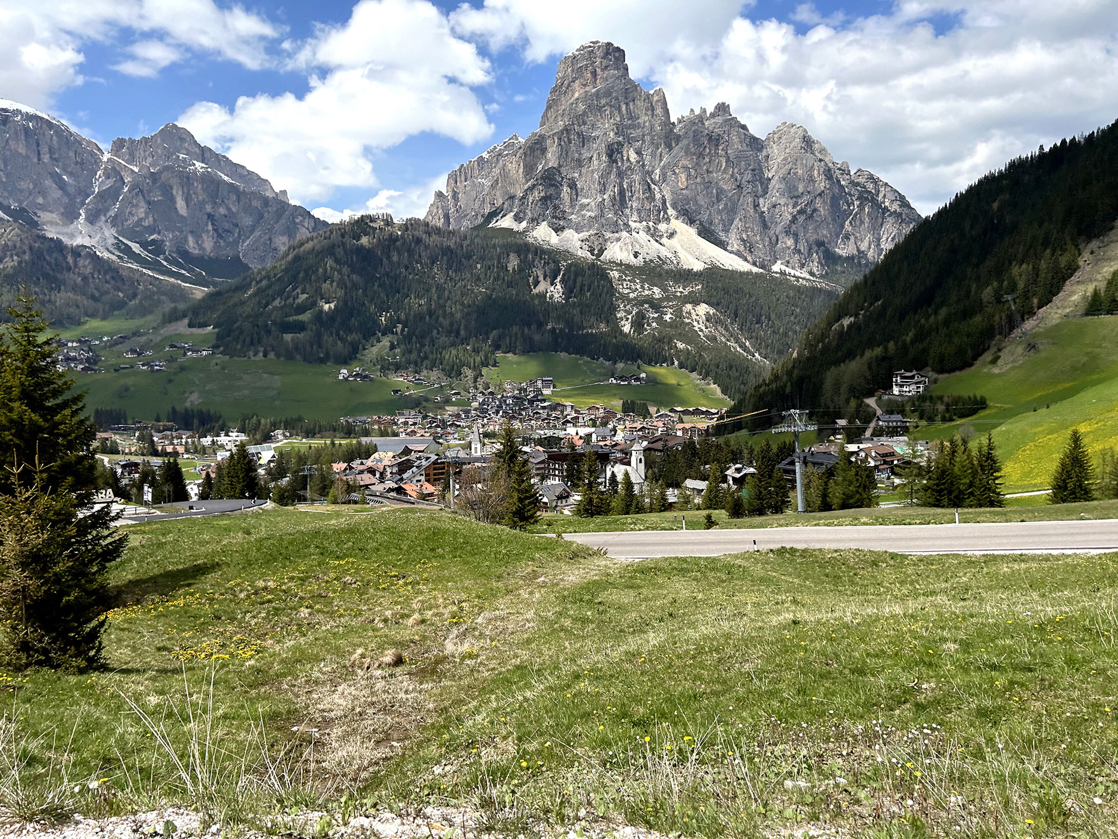 Scenic view of a mountainous landscape with a village nestled in a green valley, surrounded by towering rocky peaks under a partly cloudy sky.