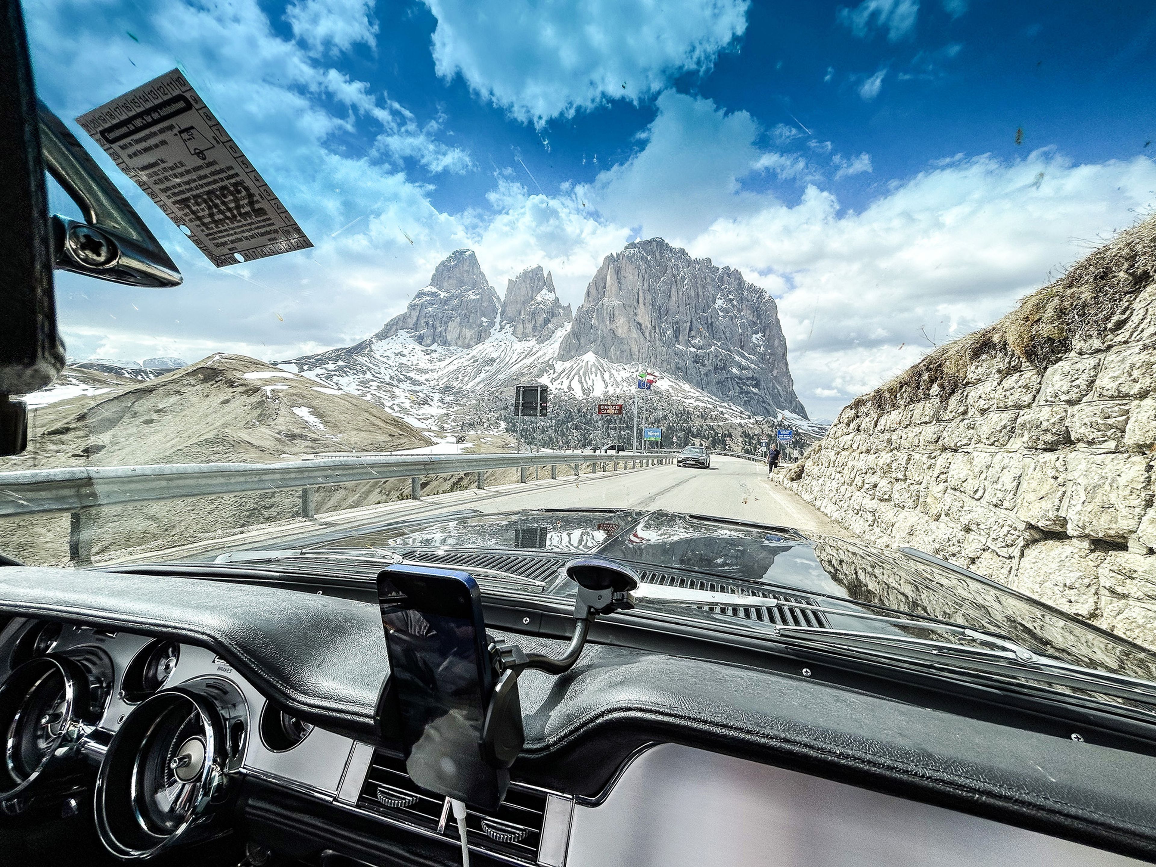 Blick aus dem Armaturenbrett eines Autos, das über eine Bergstraße mit schneebedeckten Gipfeln unter strahlend blauem Himmel fährt.
