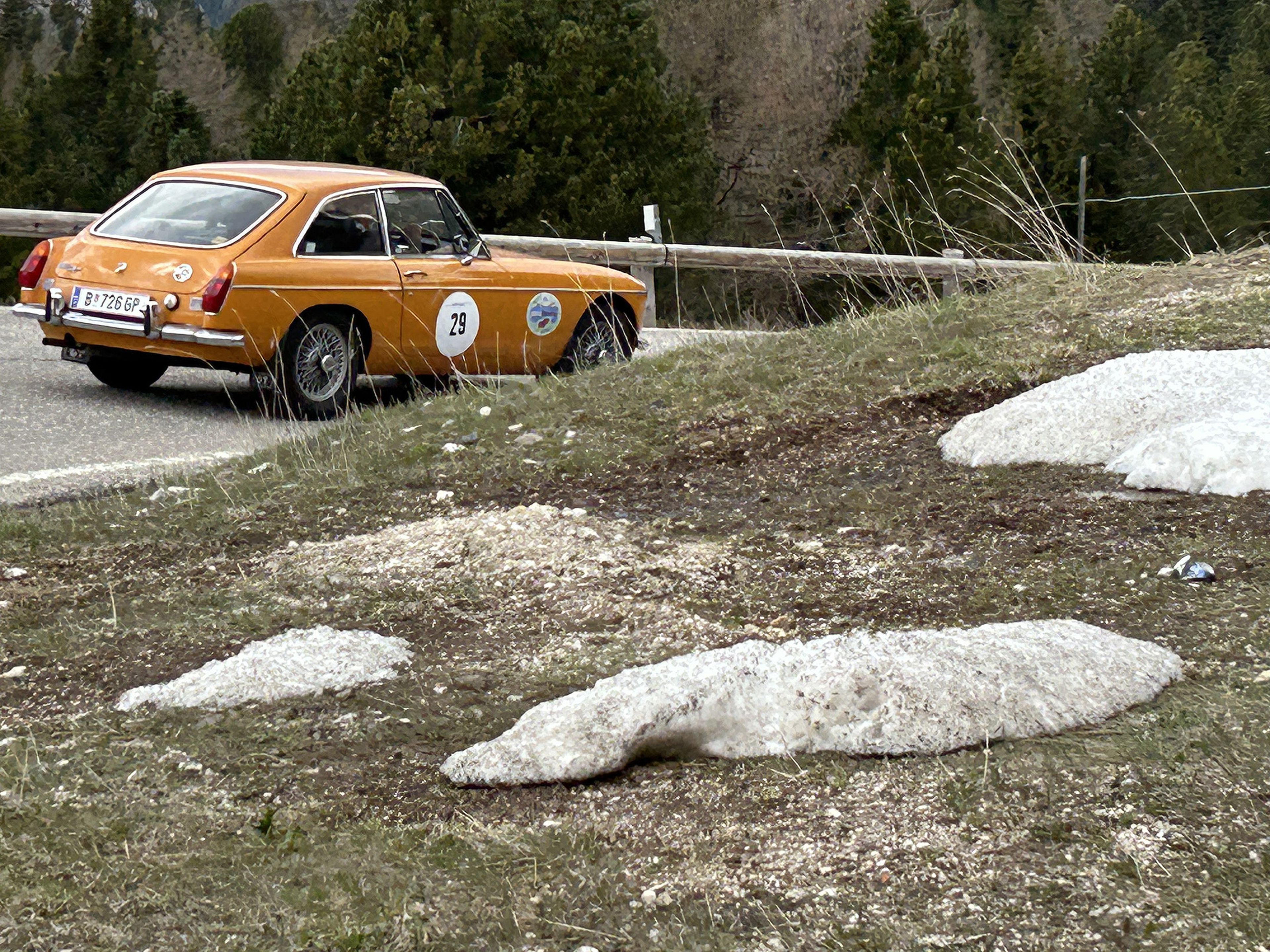 Orange vintage car with racing decals drives on a mountain road, passing patches of melting snow and surrounded by trees.
