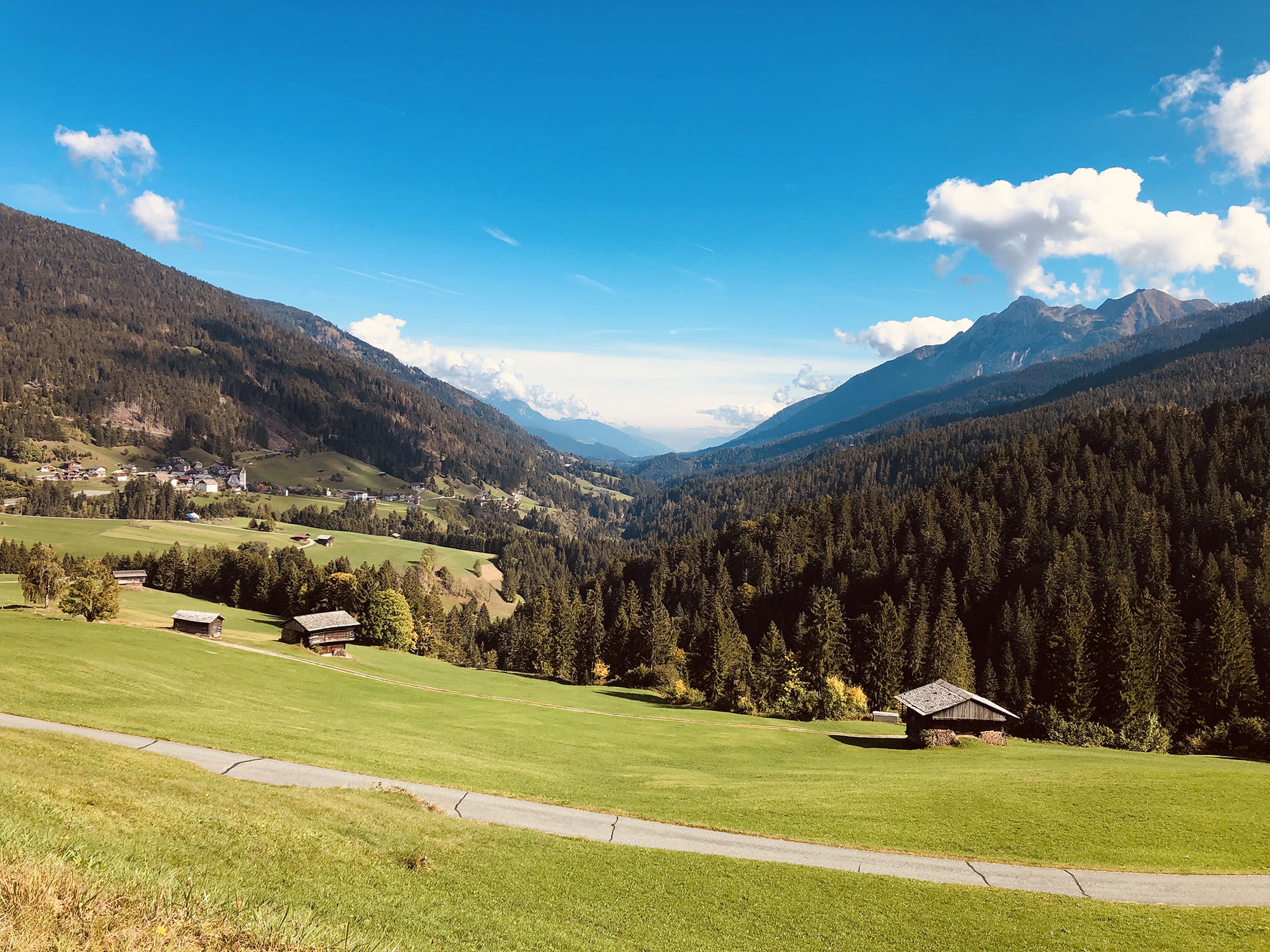 Blick auf ein üppig grünes Tal mit verstreuten Holzhütten, umgeben von bewaldeten Bergen unter einem klaren blauen Himmel mit Schäfchenwolken.