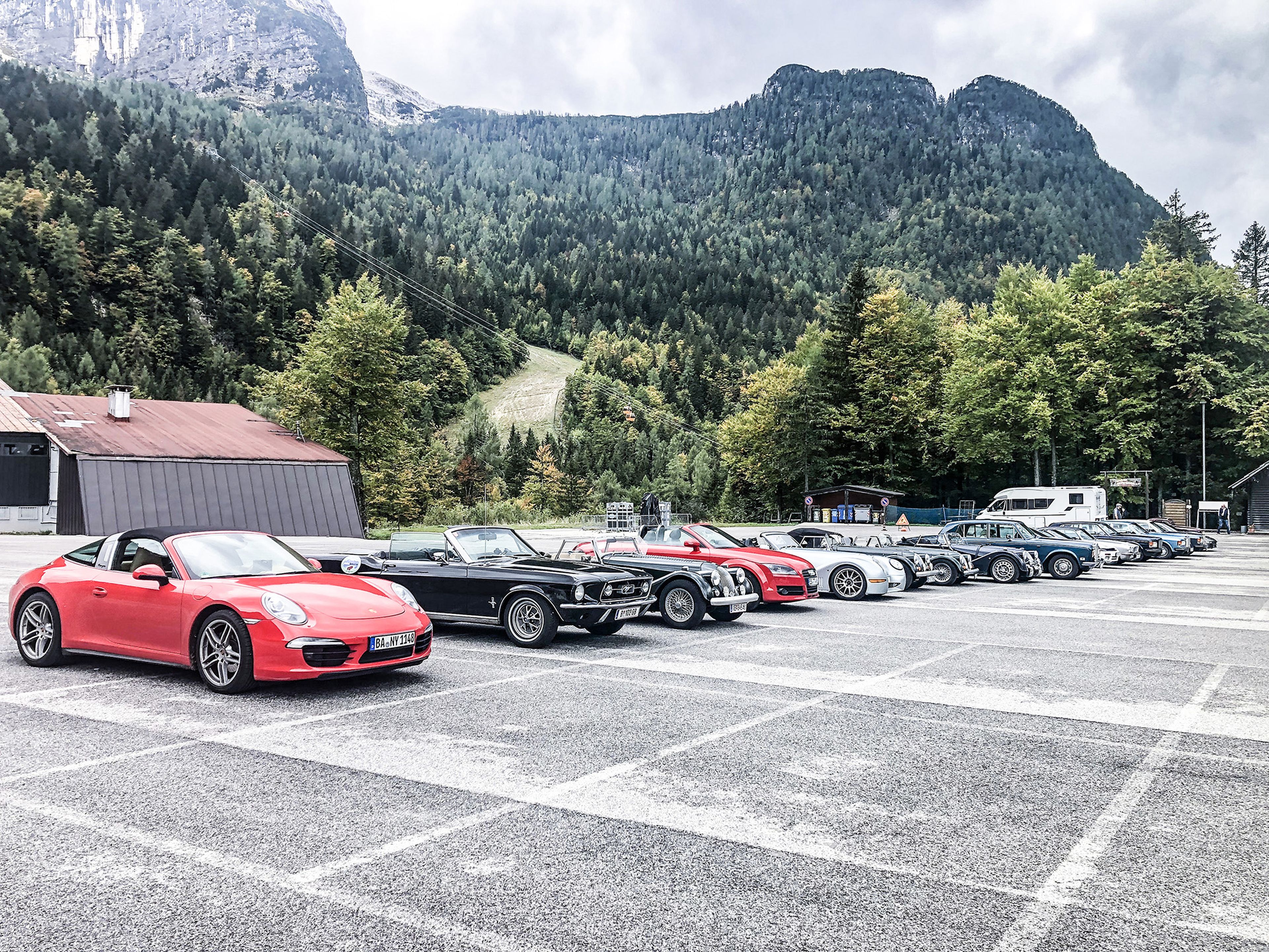 Eine Reihe verschiedener Sportwagen, die auf einem Parkplatz in den Bergen geparkt sind, umgeben von üppig grünen Bäumen und hoch aufragenden Gipfeln unter einem bewölkten Himmel.