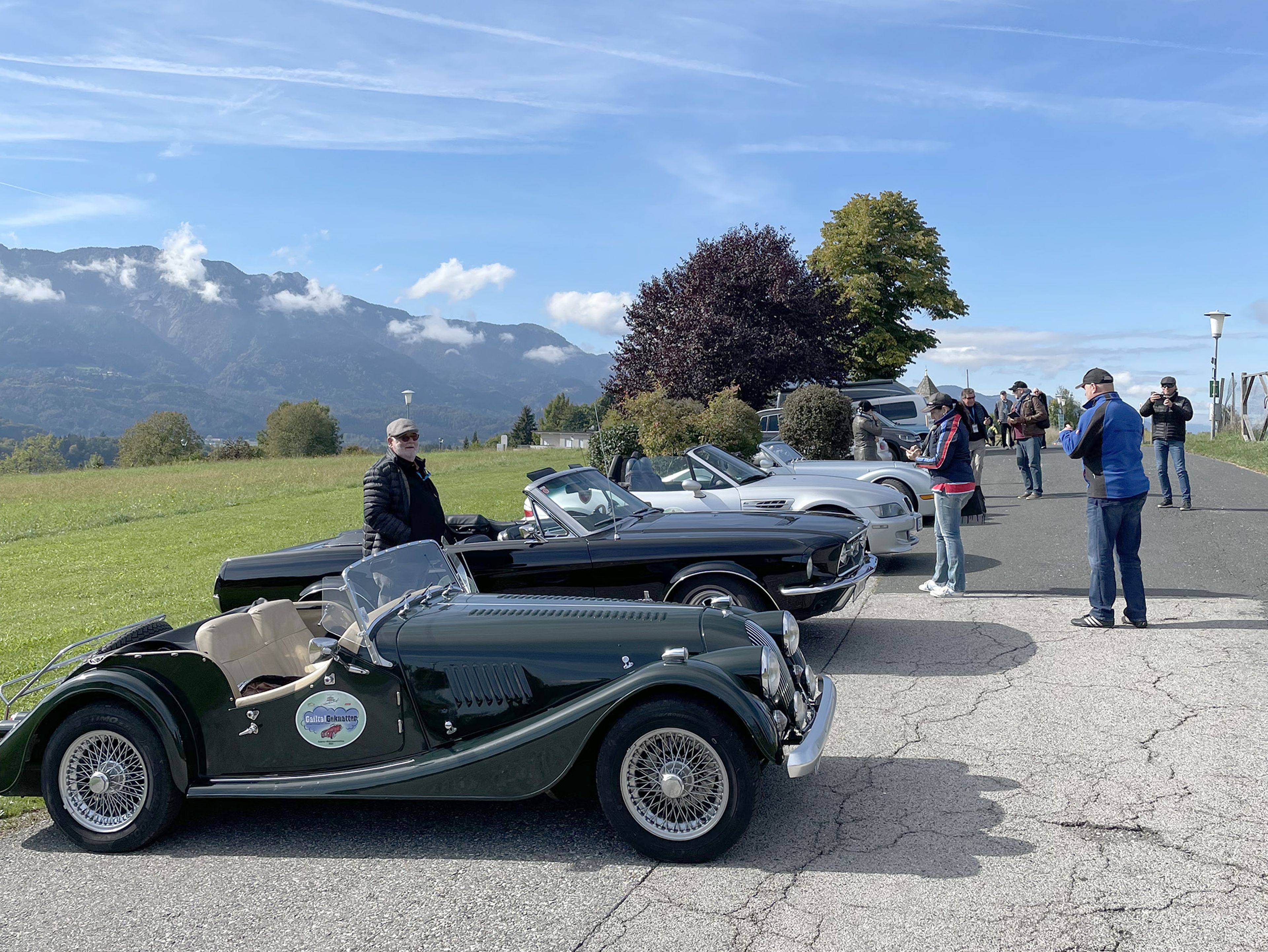 Eine Reihe von Oldtimern, geparkt auf einer malerischen Straße mit Bergen im Hintergrund. Die Leute bewundern die Autos unter einem klaren blauen Himmel.