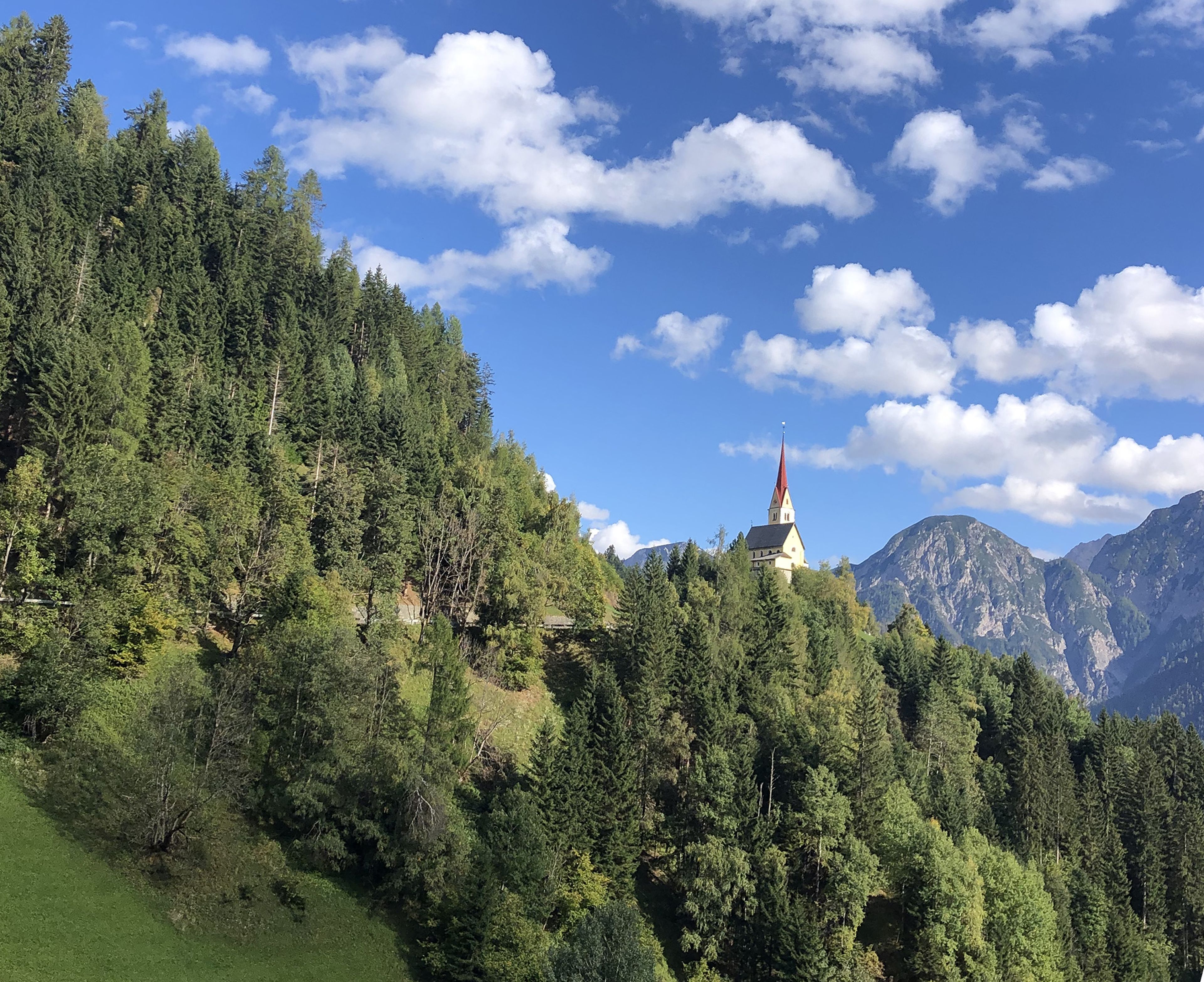 Eine kleine Kirche mit einem roten Kirchturm steht auf einem bewaldeten Hügel, umgeben von üppigem Grün und Bergen unter einem blauen Himmel mit vereinzelten Wolken.