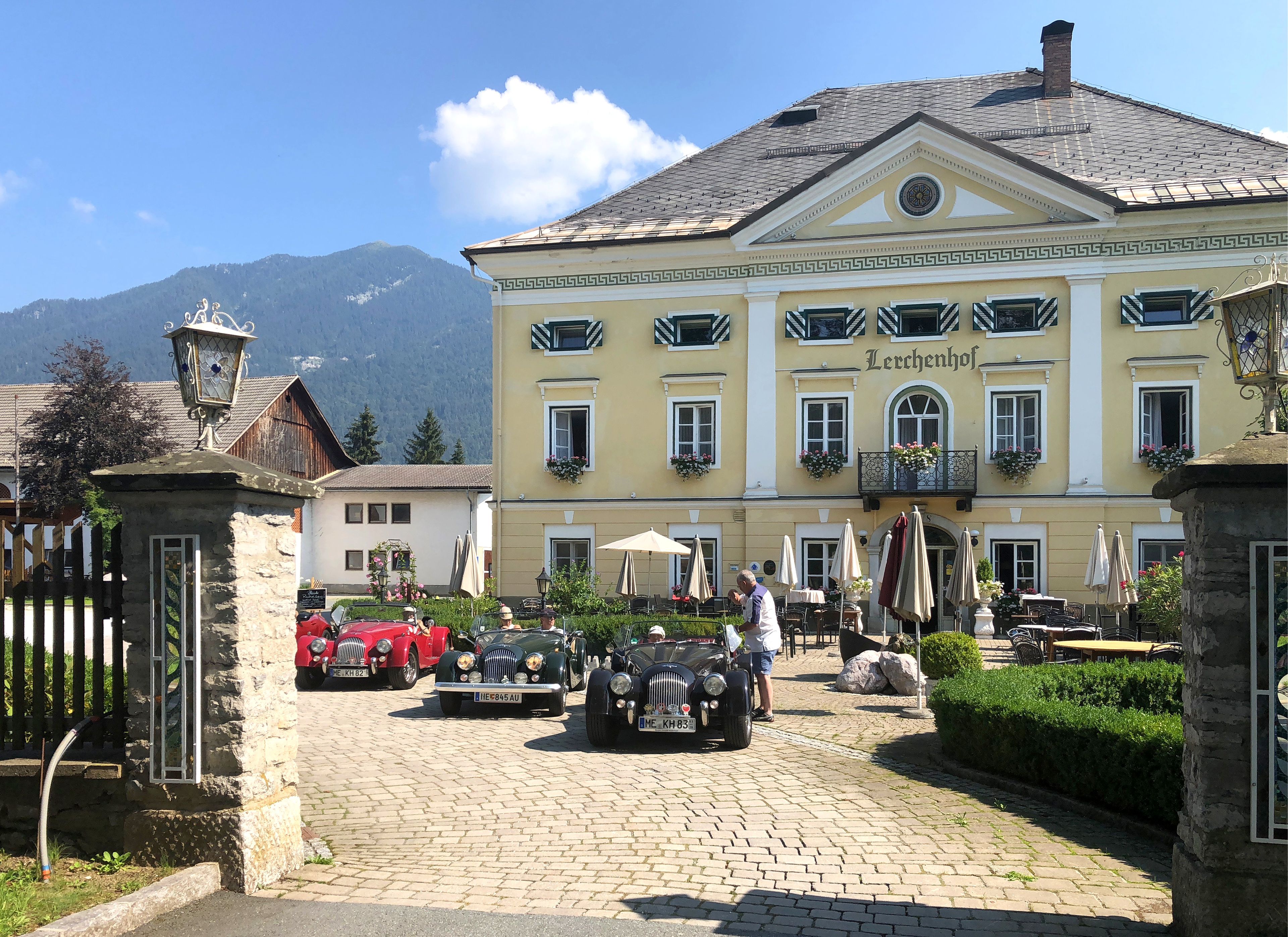 Oldtimer parken vor dem Hotel Schloss Lerchenhof mit Bergblick und klarem blauen Himmel im Hintergrund.