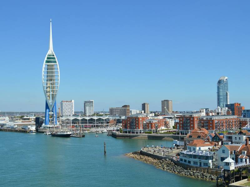 View of Portsmouth harbor featuring the Spinnaker Tower, surrounding modern buildings, and boats on a clear, sunny day.
