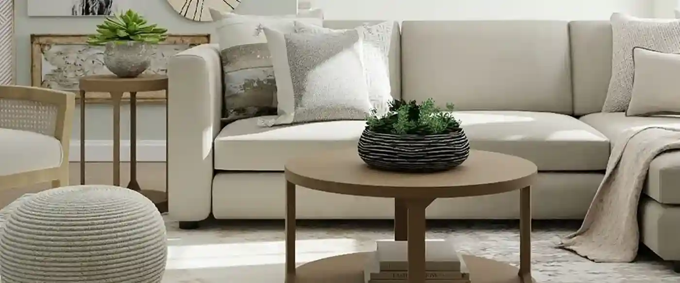 A cozy living room with a beige sectional sofa, wooden chair, round coffee table, and decorative plants, all in neutral tones.