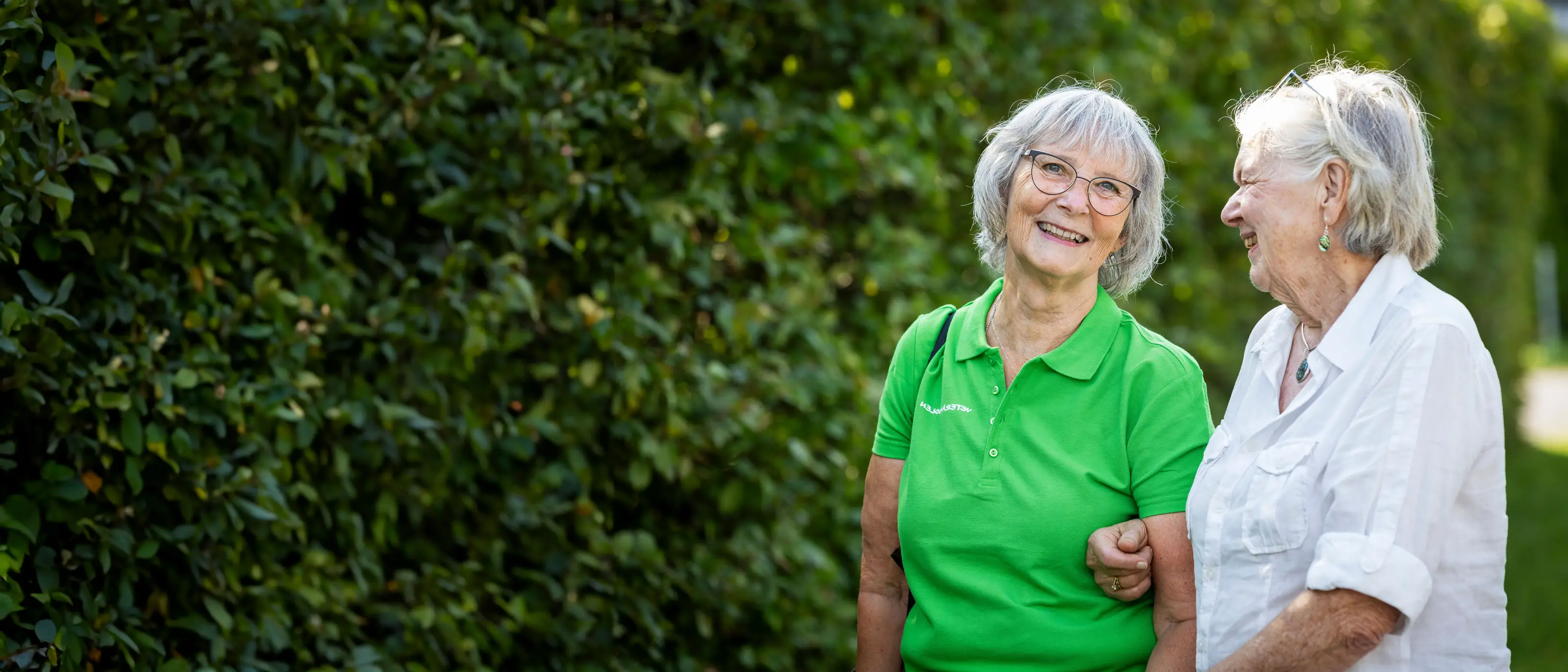 Två kvinnliga jobbonärer är ute och går i somrigt väder, de skrattar och går bredvid en grön häck