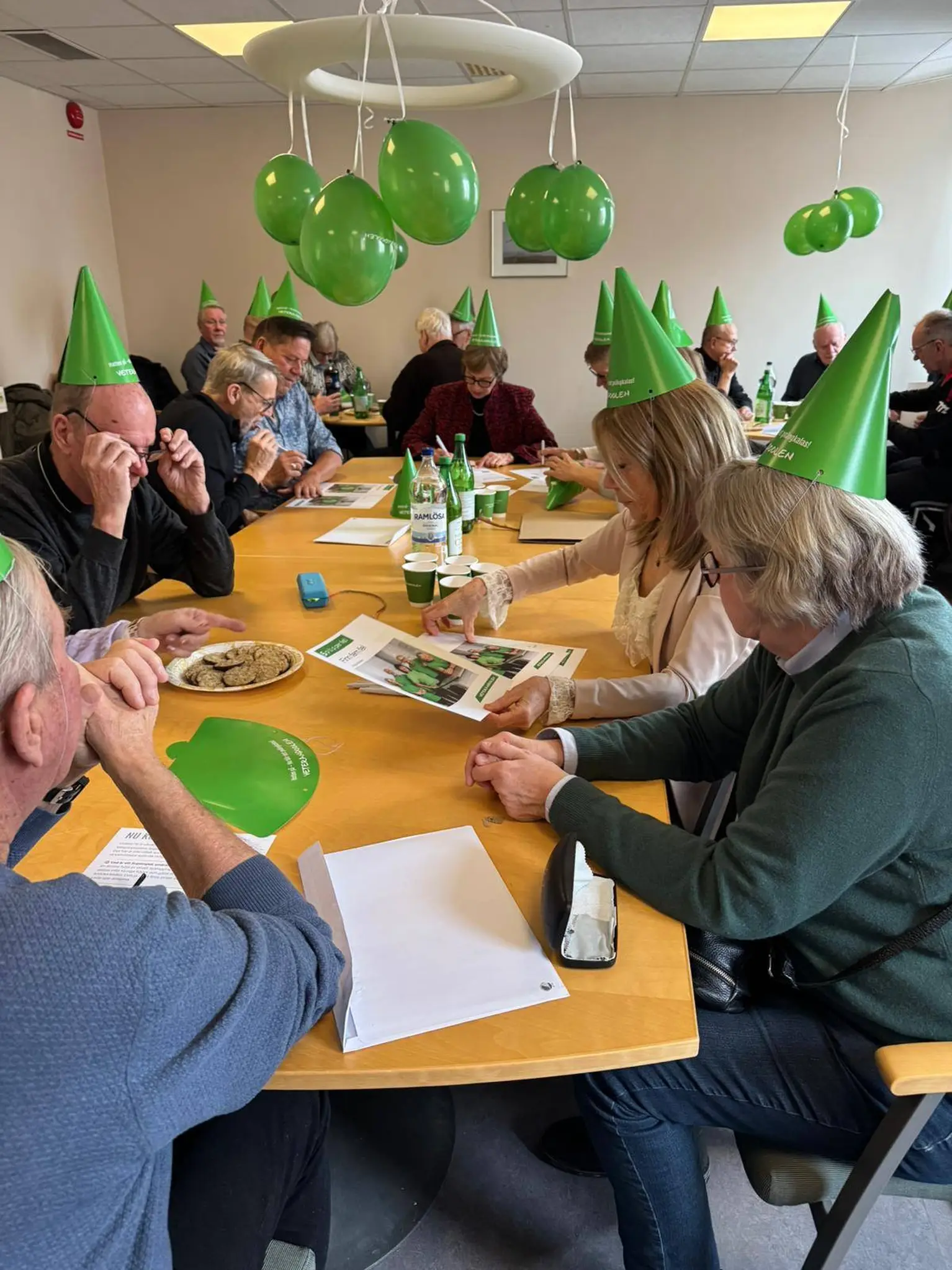 People wearing green party hats sit around a table with papers and drinks. Green balloons hang above, creating a festive atmosphere.