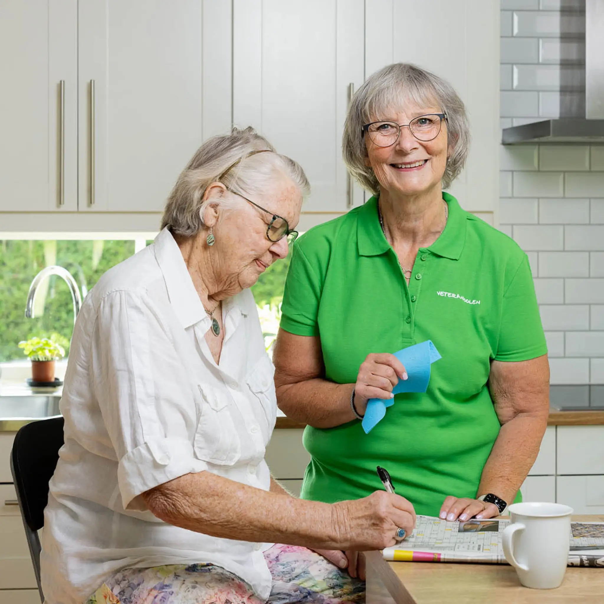 En pensionär löser korsord och en kvinnlig jobbonär står bredvid och håller en disktrasa