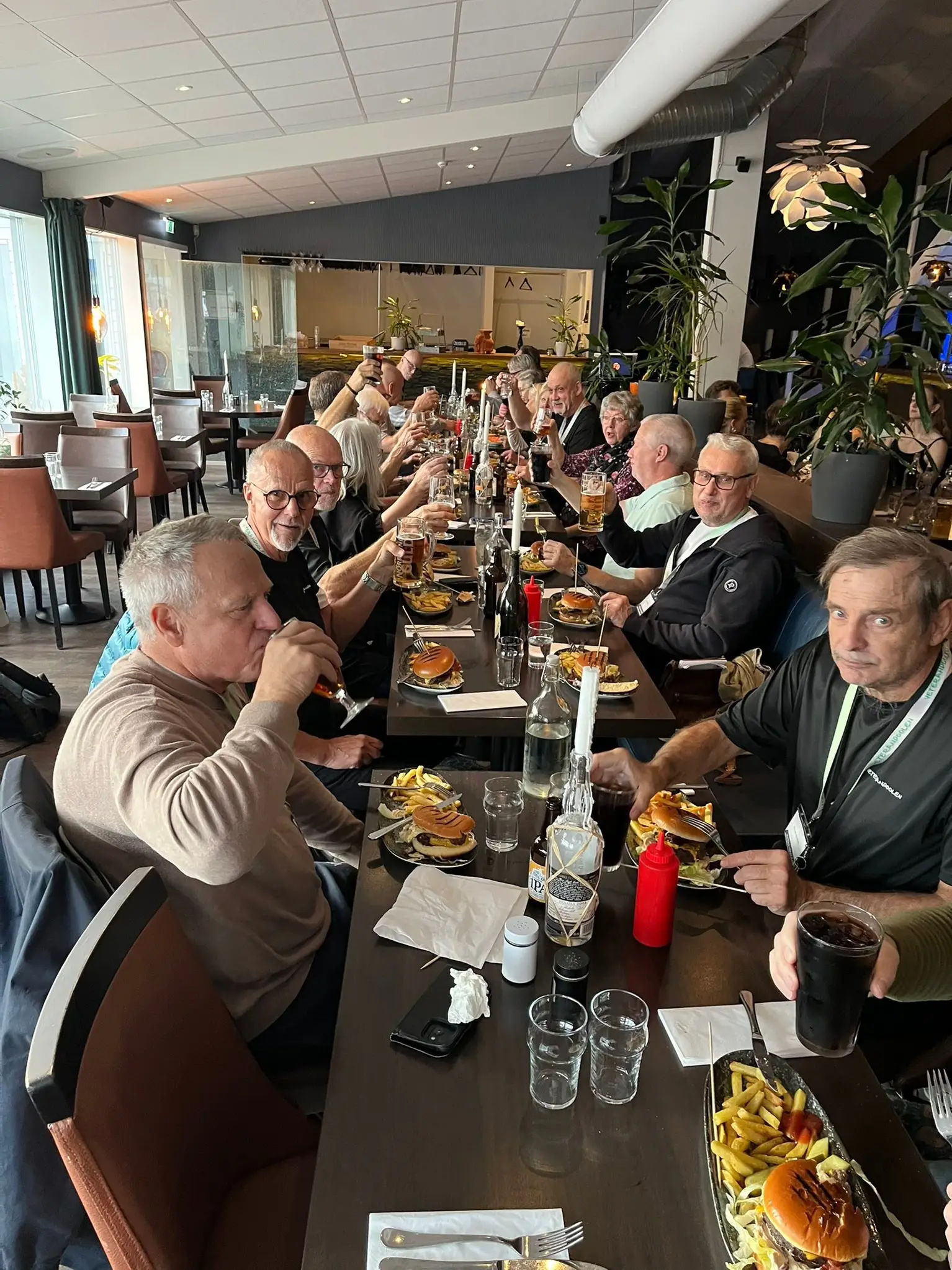 A large group of people dining at a long table in a restaurant, enjoying burgers, fries, and drinks, with glasses raised in a toast.