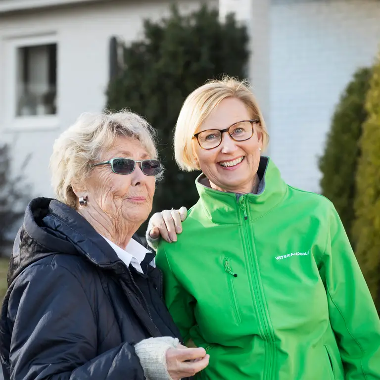 Två äldre kvinnor i går en promenad, det ser höstigt ut. Den ena kvinnan har en grön jacka och tittar mot kameran och ler.