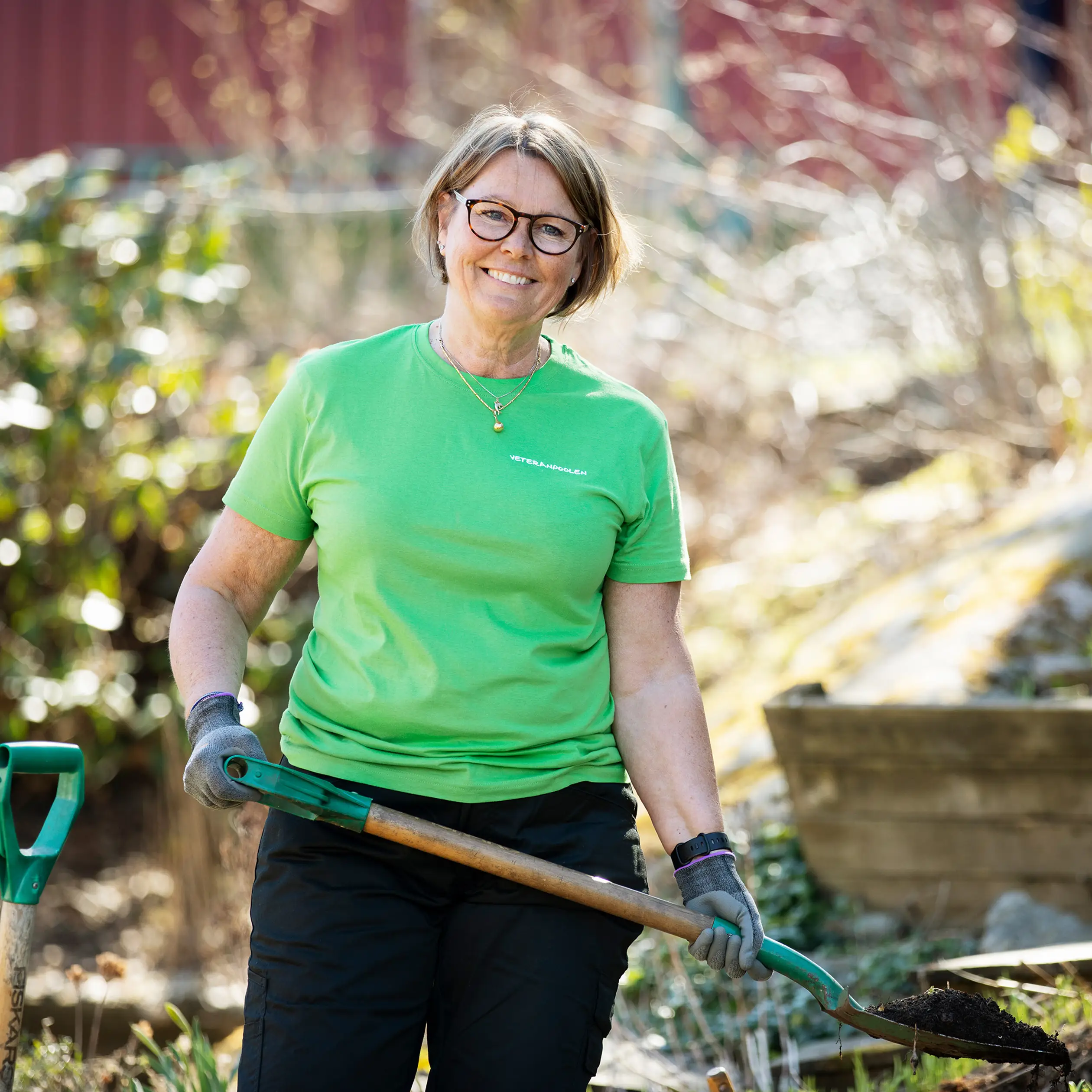 En jobbonär i grön tröja håller i en spade, hon tittar mot kameran och ler.