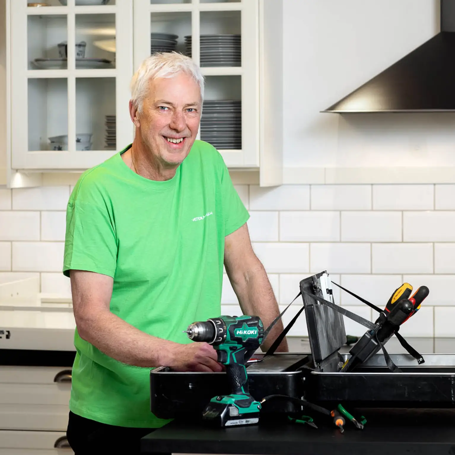 En veteran står i ett kök och har en verktygslåda, han har en grön t-shirt på sig och ler mot kameran