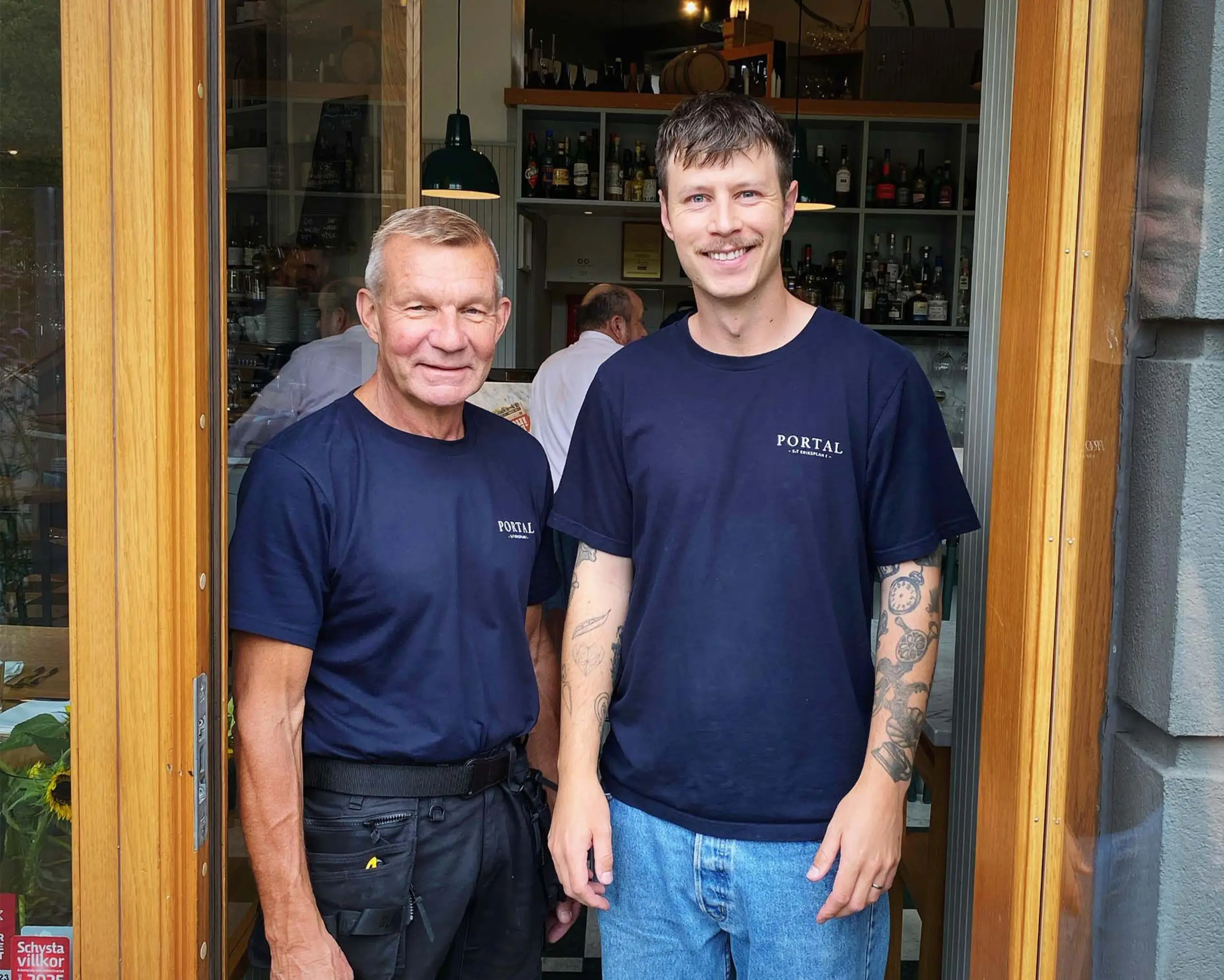Två män står i entrén till en restaurang, de har på sig mörkblå t-shirts och ler mot kameran