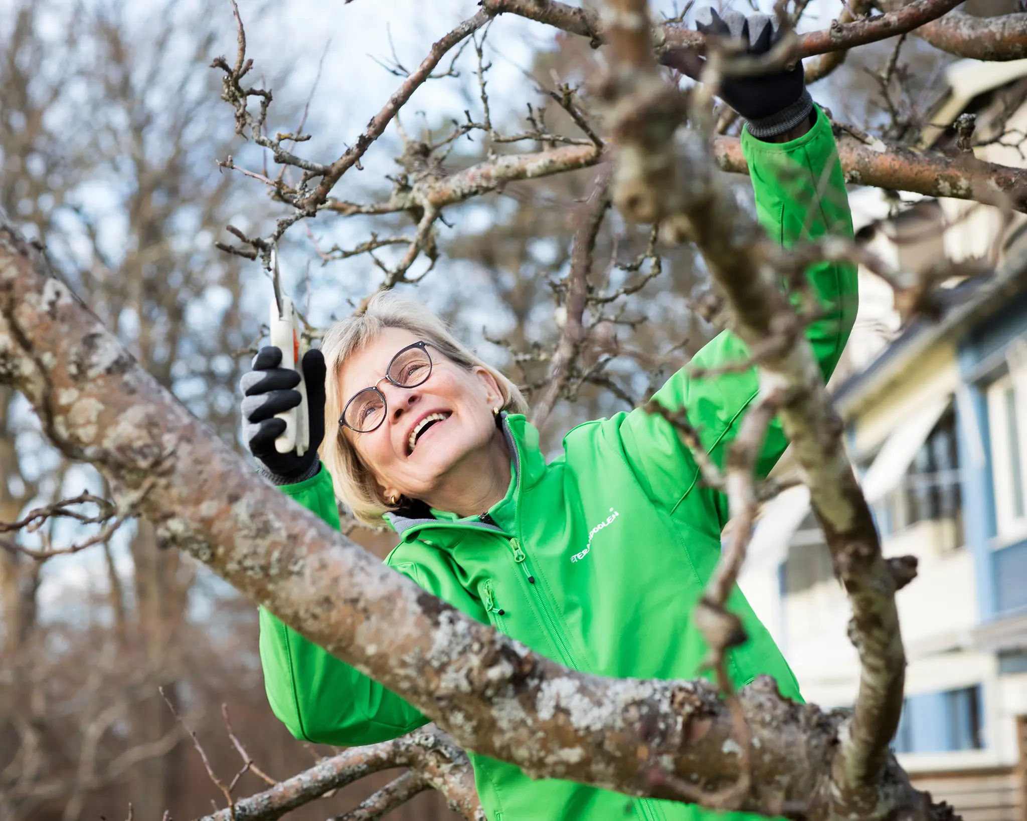 En kvinnlig veteran beskär ett träd, hon har en grön jacka på sig och ser glad ut